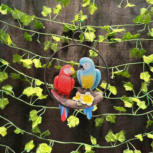 May include: Two parrots, one red and one blue, are perched on a branch inside a black metal ring. The parrots are hanging from a chain. The background is a wall covered in green ivy.